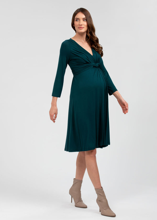 Schwangere Frau trägt enganliegendes Umstands-Strickkleid in Weiss mit grünen Streifen. Das Schwangerschaftskleid ist knielang und kurzärmelig. Es betont den Babybauch
