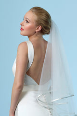 Das Model trägt einen zweilagigen Brautschleier aus weichem Soft-Tüll in Elfenbein (002 Ivory). Der Satinbesatz (0,8 cm) sorgt für eine elegante, klare Kante. Mit 75 cm Länge ist der Schleier ideal für moderne Brautlooks und lässt sich dank Haarkamm sicher in der Frisur befestigen.