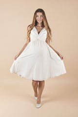 Das Model trägt das Umstands-Hochzeitskleid Ely in Off-White. Der V-Ausschnitt und das weich drapierte Oberteil wirken elegant, der knielange Rock fällt fliessend und bietet dem Babybauch angenehm Platz – ideal für Ziviltrauung/Standesamt.