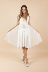 Das Model trägt das kurze Umstands-Hochzeitskleid Fiona in schlichtem Weiss. Der V-Ausschnitt und das figurbetonte Oberteil wirken elegant, während der luftige, weit schwingende Rock dem Babybauch angenehm Platz gibt – ideal für Ziviltrauung/Standesamt und eine moderne, unkomplizierte Hochzeit.