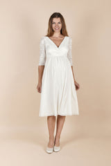 Das Model trägt das Umstands-Hochzeitskleid Cecilia in Off-White in einem warmen Indoor-Setting. Das Kleid hat ein Spitzenoberteil mit V-Ausschnitt und 3/4-Ärmeln sowie einen weich fliessenden Rock, der dem Babybauch angenehm Platz gibt – ideal für Ziviltrauung/Standesamt. Hinweis: Schärpe nicht enthalten (separat erhältlich).