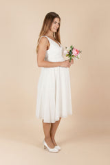 Seitenansicht des kurzen Umstandskleids Larina in Weiss: Das ärmellose Oberteil mit V-Ausschnitt wirkt elegant, der weich fliessende Rock bietet dem Babybauch angenehm Platz. Ein stilvoller Look für Standesamt, Sommerfeste oder besondere Anlässe in der Schwangerschaft.