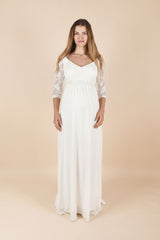Das Model trägt das Umstands-Hochzeitskleid Vivien in Weiss. Das Kleid wirkt schlicht und elegant, mit ärmelloser/oberer Partie und zarten, transparenten Ärmeln sowie einem lang fliessenden Rock, der dem Babybauch angenehm Platz gibt – ideal für Ziviltrauung/Standesamt.