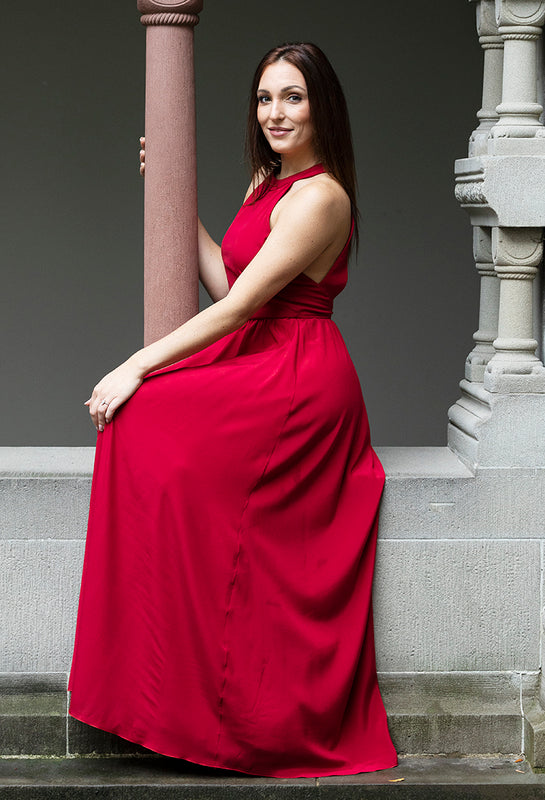 Frau sitzt in elegantem, langem, rotem Seiden-Abendkleid, welches schulterfrei ist.