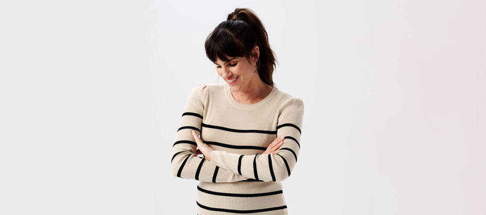 Schwangere Frau trägt einen Umstands-Pullover in beige mit schwarzen Streifen. Der Pullover hat lange Puffärmel.