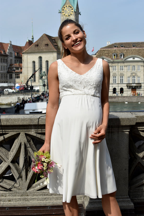 Schwangere Frau trägt ärmelloses Umstands-Hochzeitskleid mit V-Ausschnitt und Spitzenoberteil.