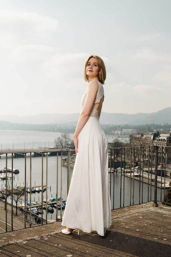 Schlanke Frau trägt langes Hochzeitskleid mit Spitzen-Cutout-Rücken. Das elegante und schmal geschnittene Kleid eignet sich für Standesamt und Kirche. Im Hintergrund sieht man den Zürichsee.